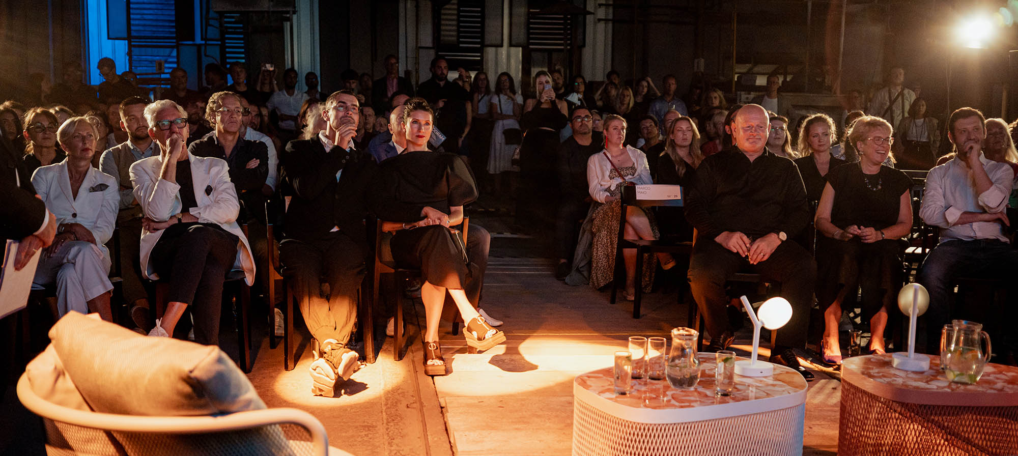 A stylish audience sits in moody light, faces aglow from sculptural handblown crystal lamps; glasses shimmer on chic tables.
