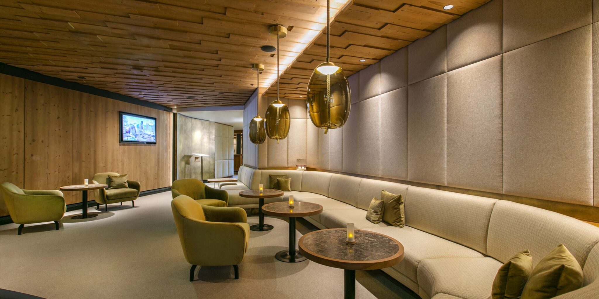 A modern lounge with beige padded walls, cream sofa, Brokis Knot Uovo pendants, dark tables, and plush green armchairs.