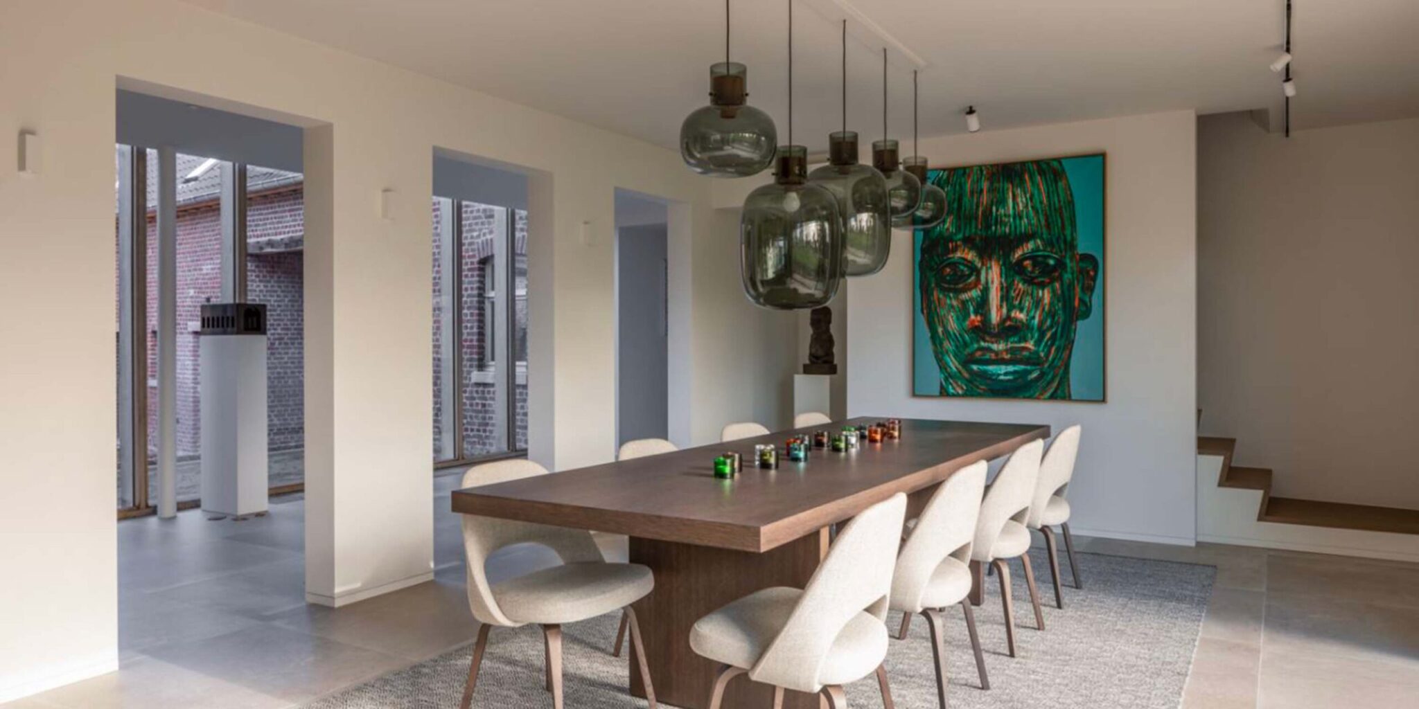 Modern dining room with a long wooden table, white chairs, Brokis Awa pendants, and bold portrait art.
