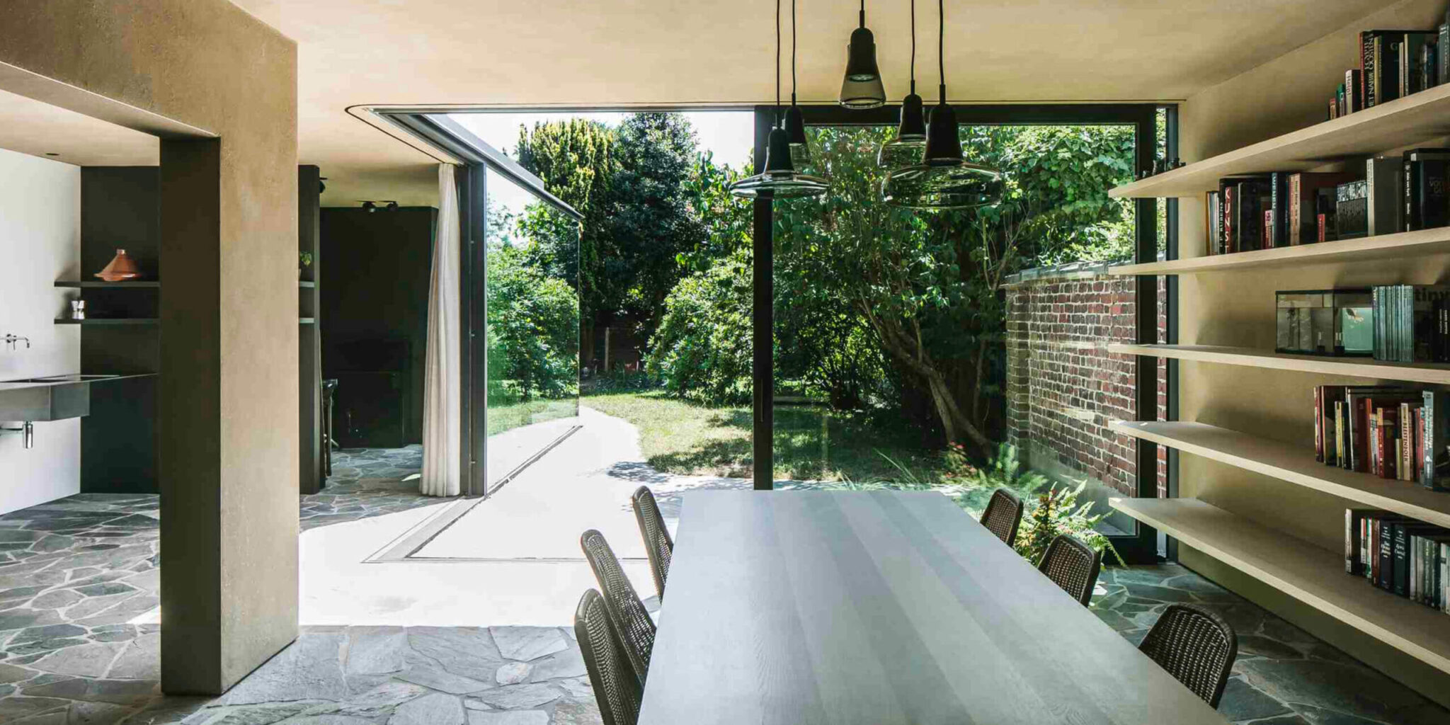 Modern dining area with long table, woven chairs, open bookshelves, stone floors, and Brokis Shadows Set Free.