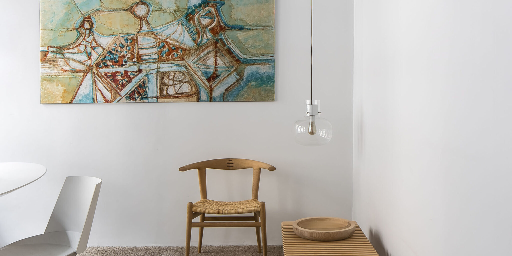 A minimalist room with a wooden chair, slatted bench, round bowl, Brokis Awa Small pendant, and abstract blue art.