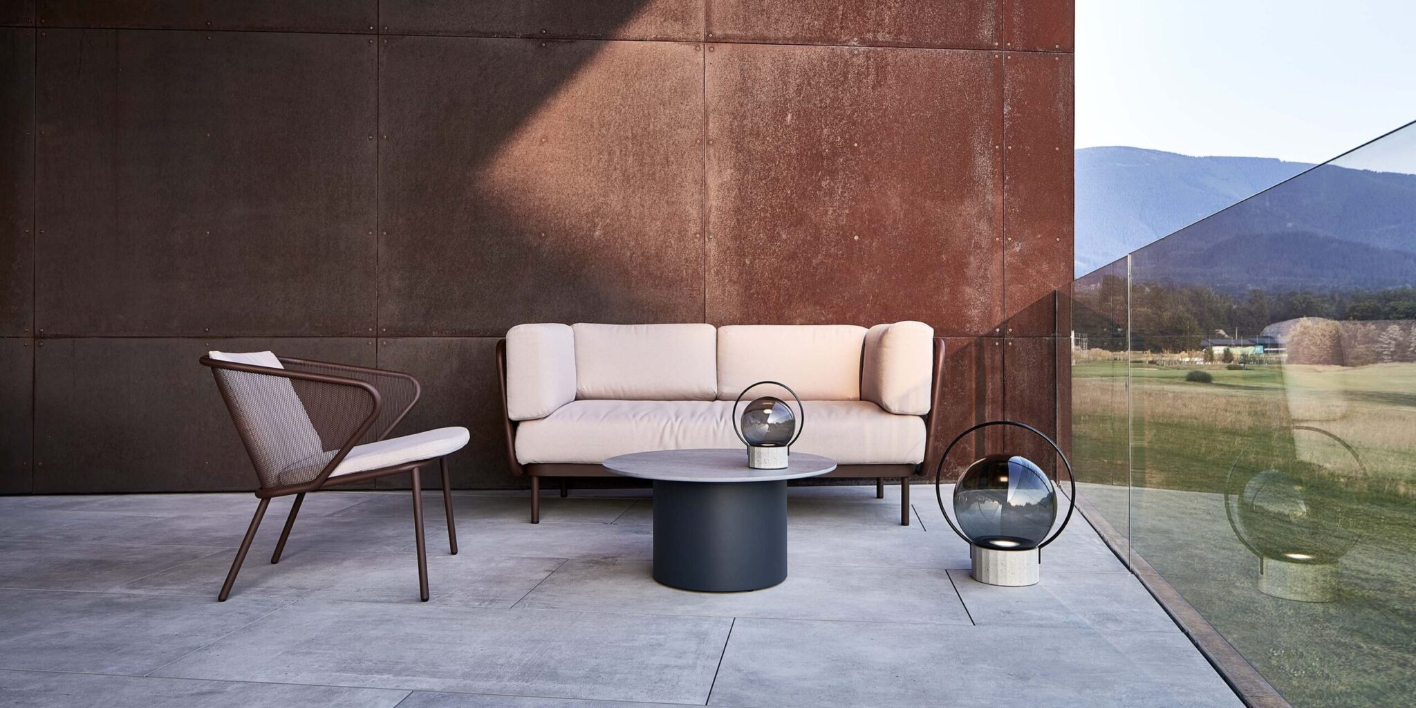 Chic patio with tan cushioned sofa, mesh chair, and Brokis Sfera Portable lanterns on a sleek grey table.
