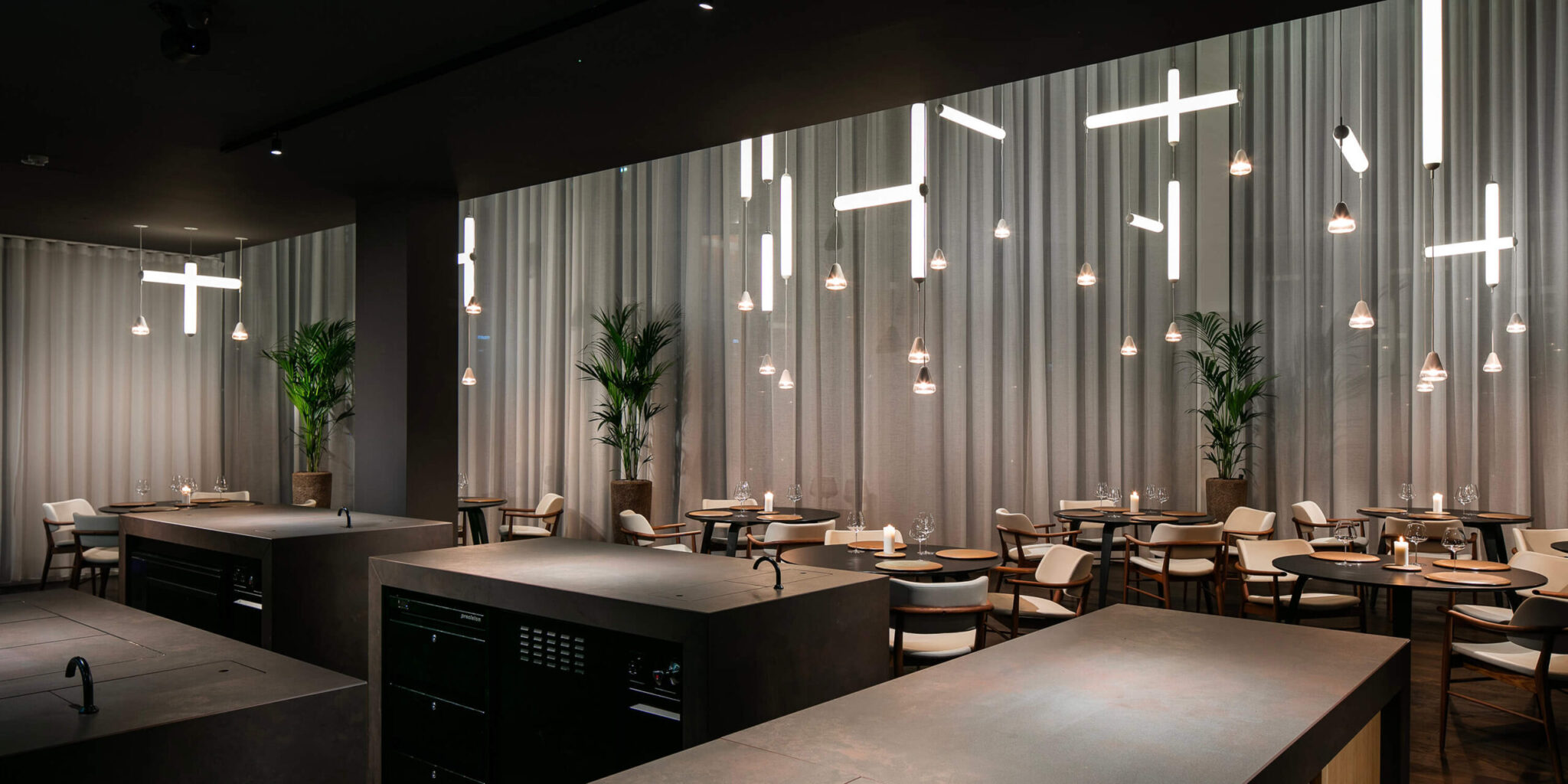 Chic restaurant with round tables, cream chairs, lush plants, sheer curtains, and Brokis Puro pendant lights above.
