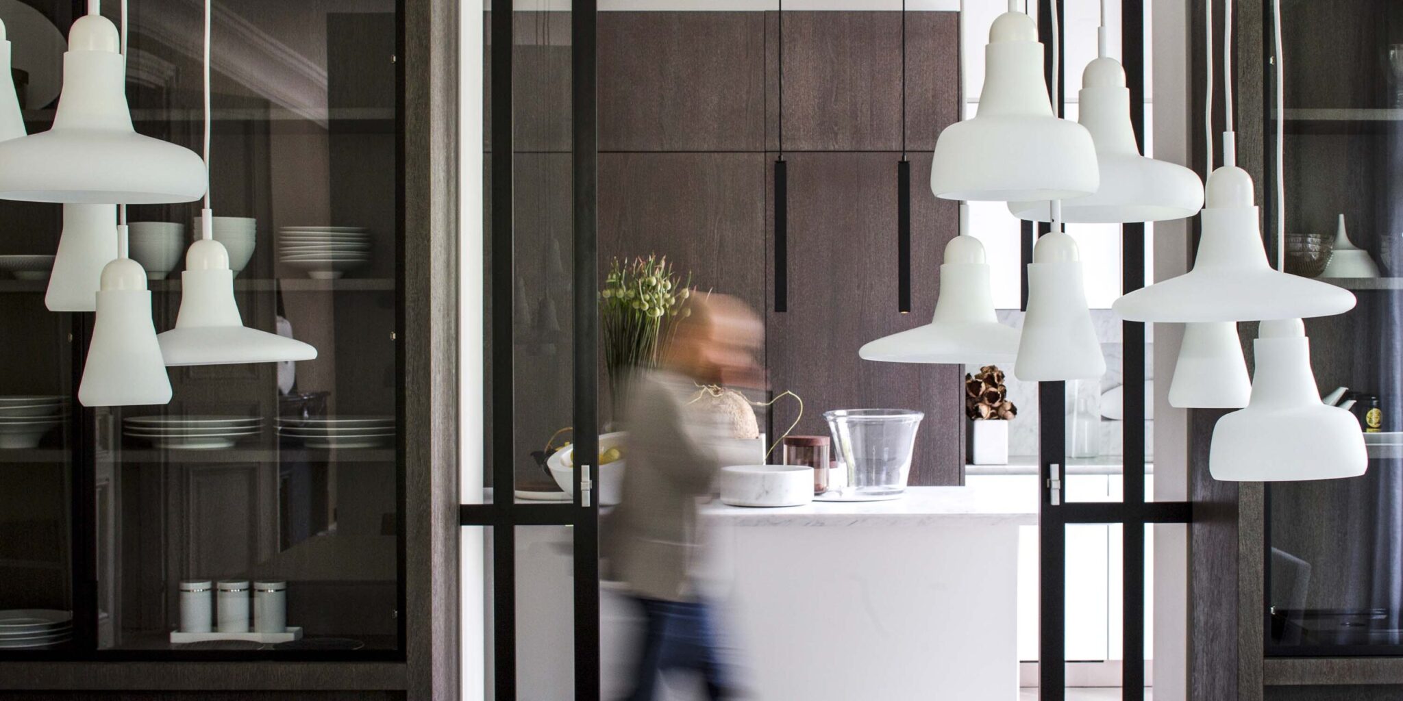 A blurred figure passes through a sleek kitchen featuring dark cabinetry, white worktops, and Brokis Shadows pendants.