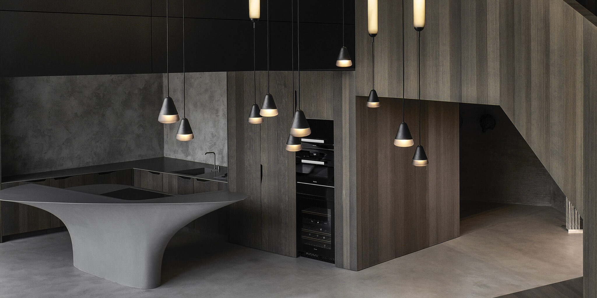 A modern kitchen with dark wood cabinetry, sculptural grey island, and Brokis Puro pendants exuding quiet luxury.