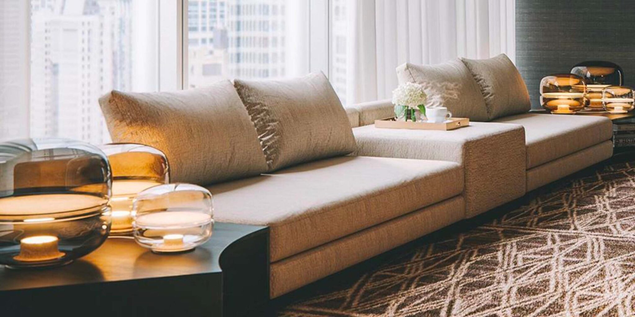 A modern beige corner sofa sits by city-view windows, flanked by Brokis Macaroon lamps and elegant décor accents.