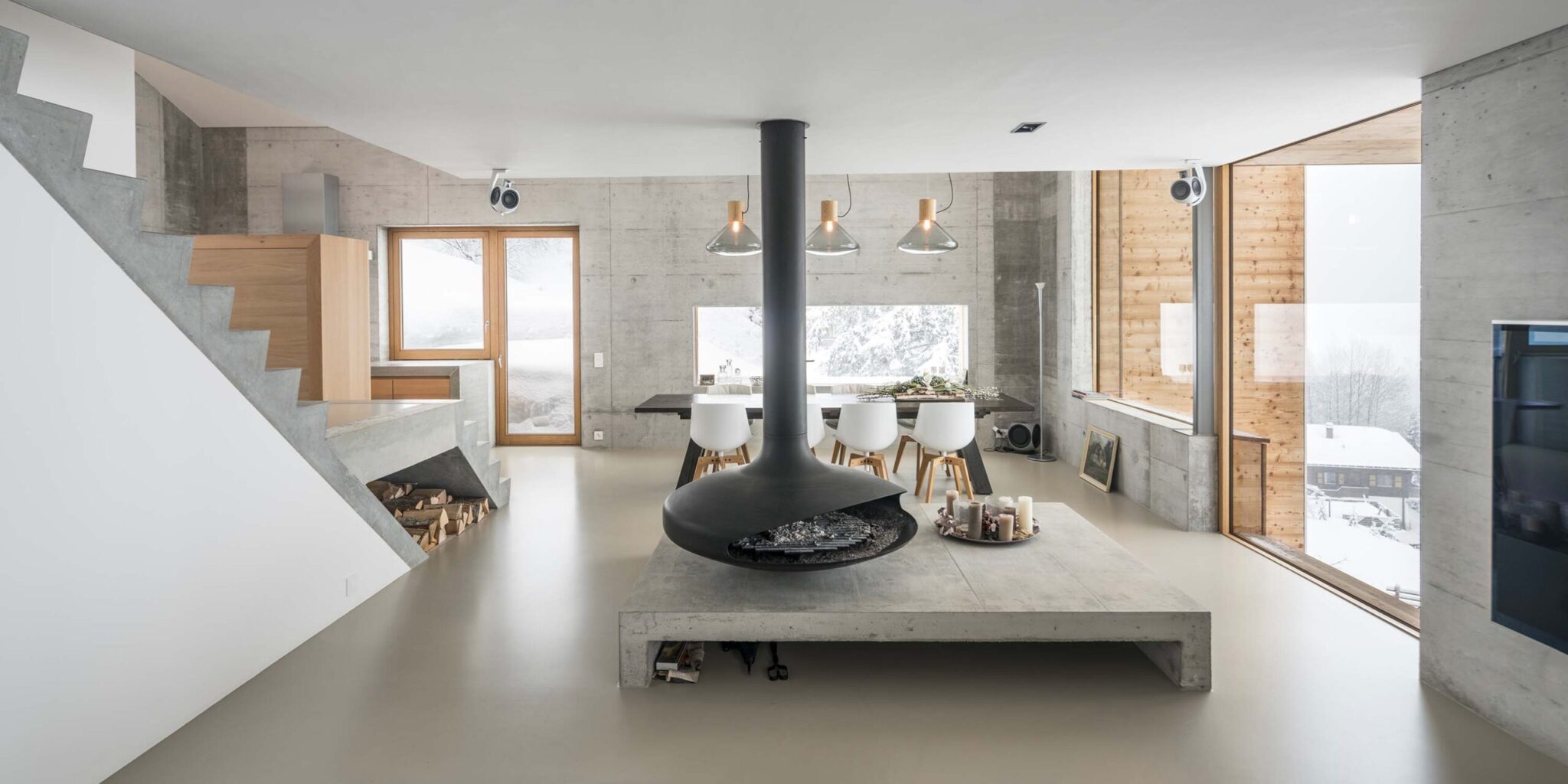 A modern, minimalist living-dining space with suspended fireplace, snowy views, Brokis Muffins, concrete and sleek stairs.