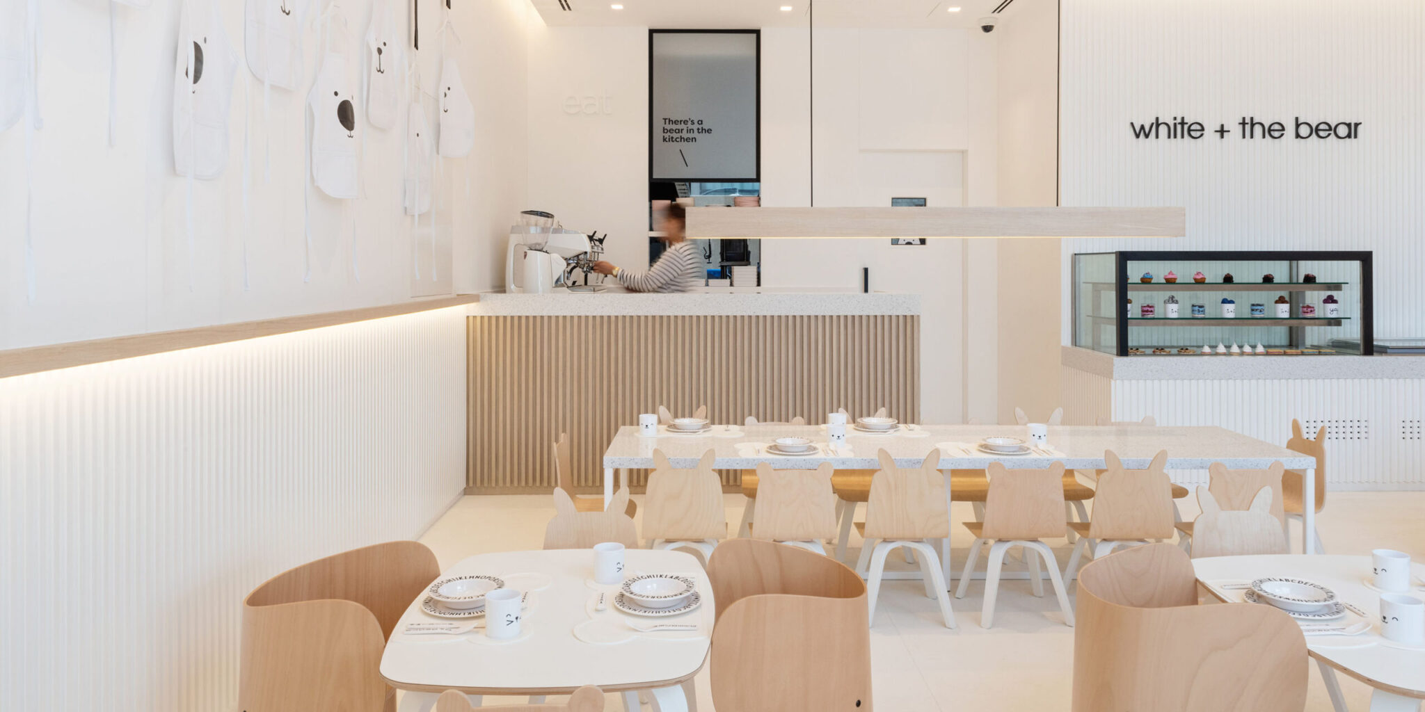 A modern, minimalist café with white walls, light wood furniture, playful chairs, elegant pendant lights, and laid tables.