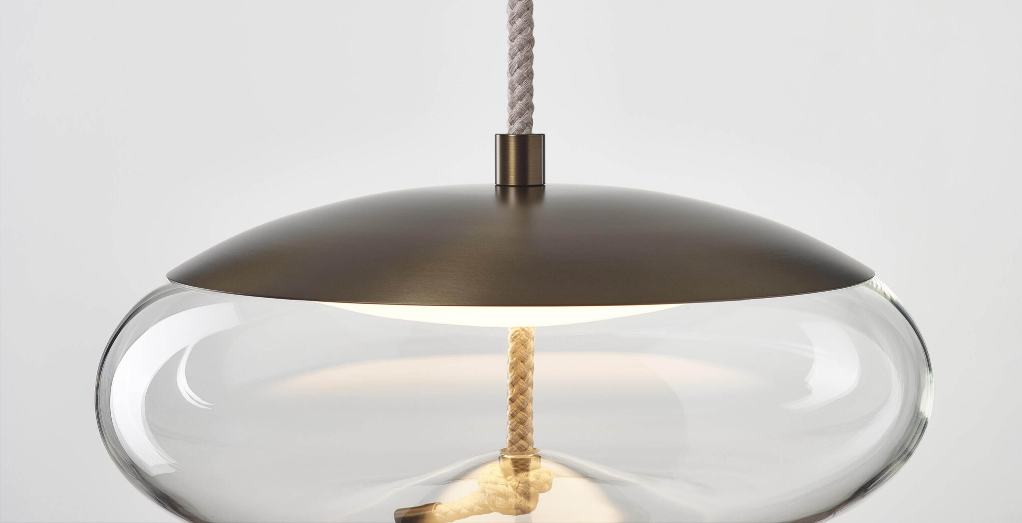 A Brokis Knot Disco pendant light with a metal dome, crystal-clear glass ring, and rope, aglow against a white background.