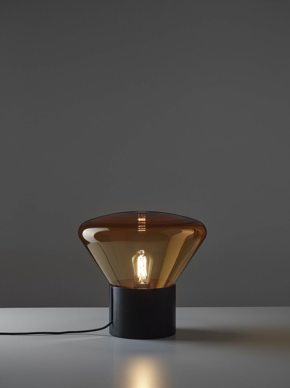 Brokis Muffins Wood 01 lamp with a handblown violet glass and black oak base glows softly on a white surface.