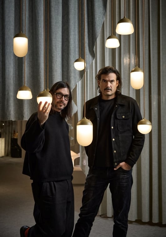 VRTIŠKA & ŽÁK stand among Brokis Planets MINI with warm bulbs, framed by tall, textured curtains.