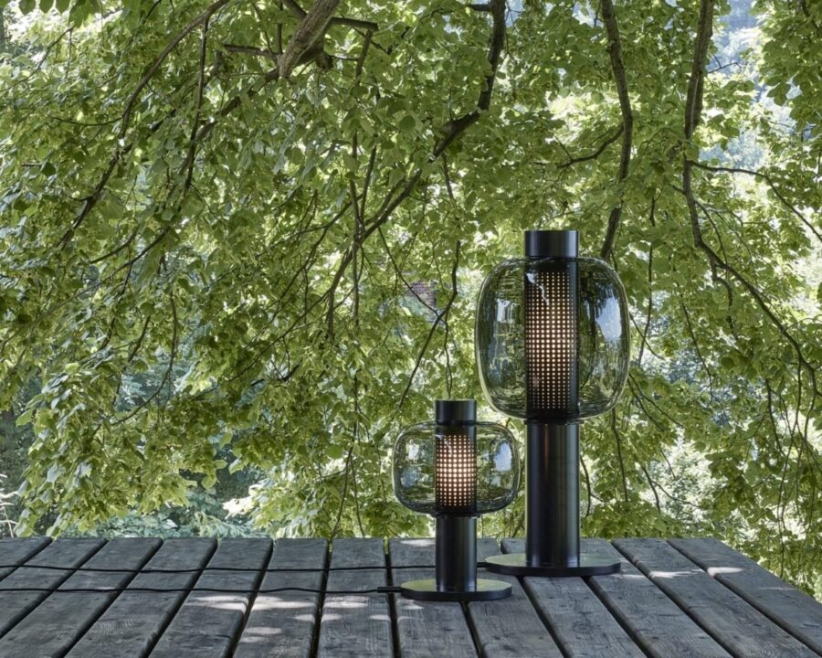 Sustainable Brokis lamps in dark metal rest on a wooden deck, their sculptural glass softly mirroring dappled sunlight.