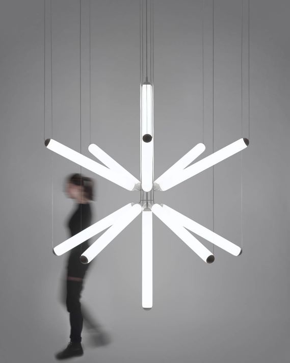 A blurred figure passes a Brokis Puro Sparkle chandelier, tubular crystal lights radiating in starburst elegance on grey.