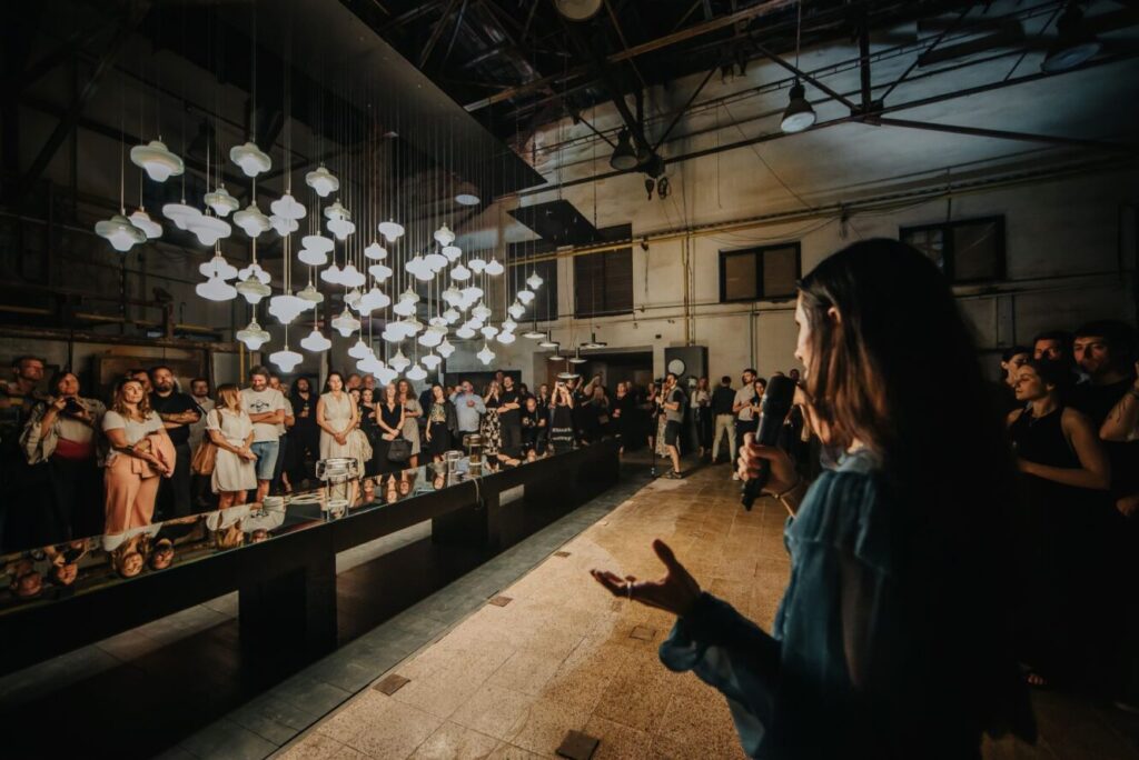 Lucie Koldova addresses an attentive audience in an industrial space, Brokis lights gleaming above a sleek reflective table.
