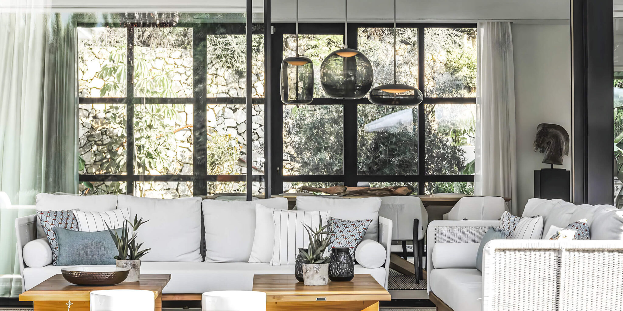 Modern living room with glass walls, white sofas, patterned cushions, wooden tables with plants, and Brokis Knot pendants.