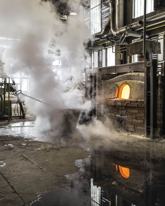 A worker tends a glowing Brokis furnace as white steam rises, softening the scene reflected on the wet factory floor.