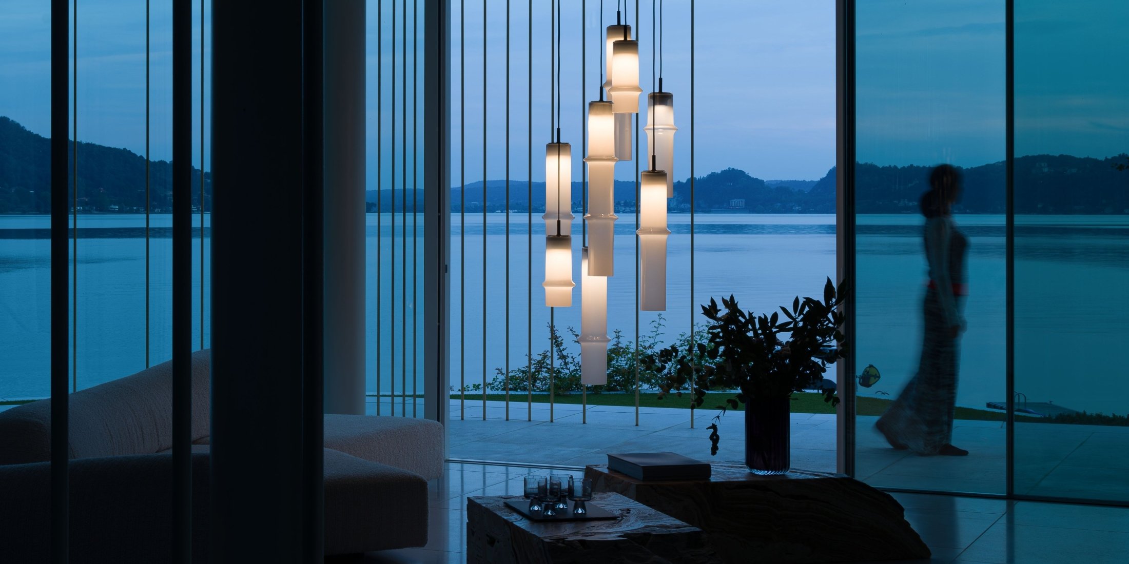 Brokis Bamboo Forest pendants glows in a modern living room at dusk, framing lake views through expansive glass walls.
