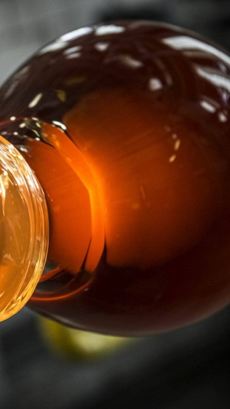 Molten liquid cascades from a handblown Brokis glass, luminous against a dark, blurred background.