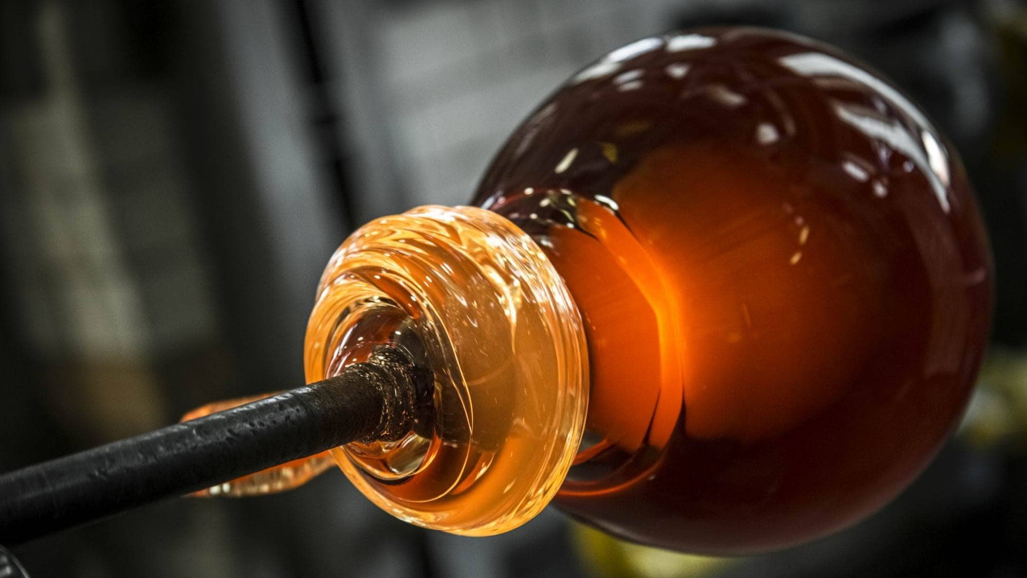 Brokis molten glass glows orange, handblown on a blowpipe; glossy and luminous against a dark, workshop backdrop.
