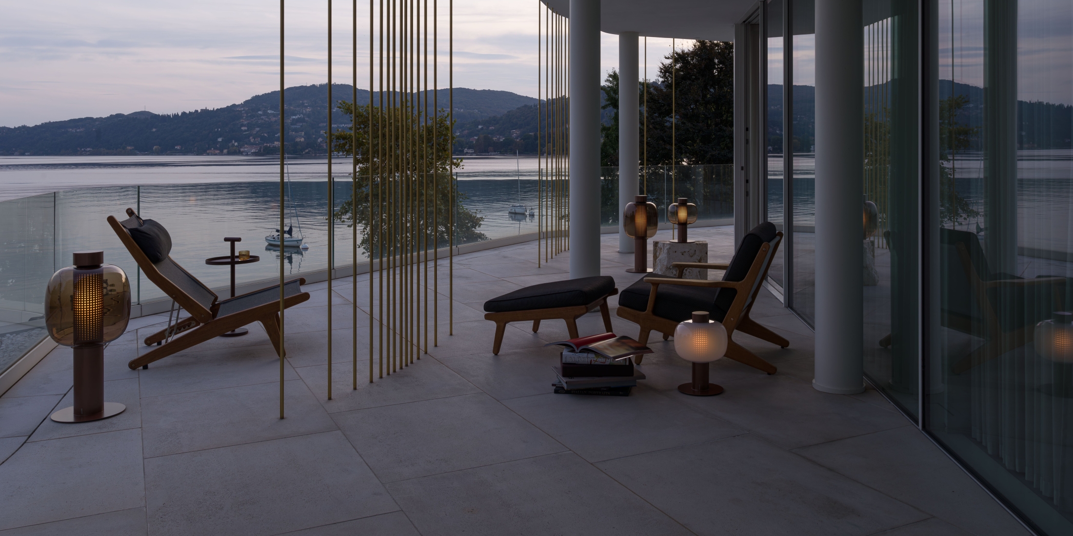 Brokis Bonbori outdoor lanterns on a modern patio with glass walls overlooks a tranquil lake at dusk.