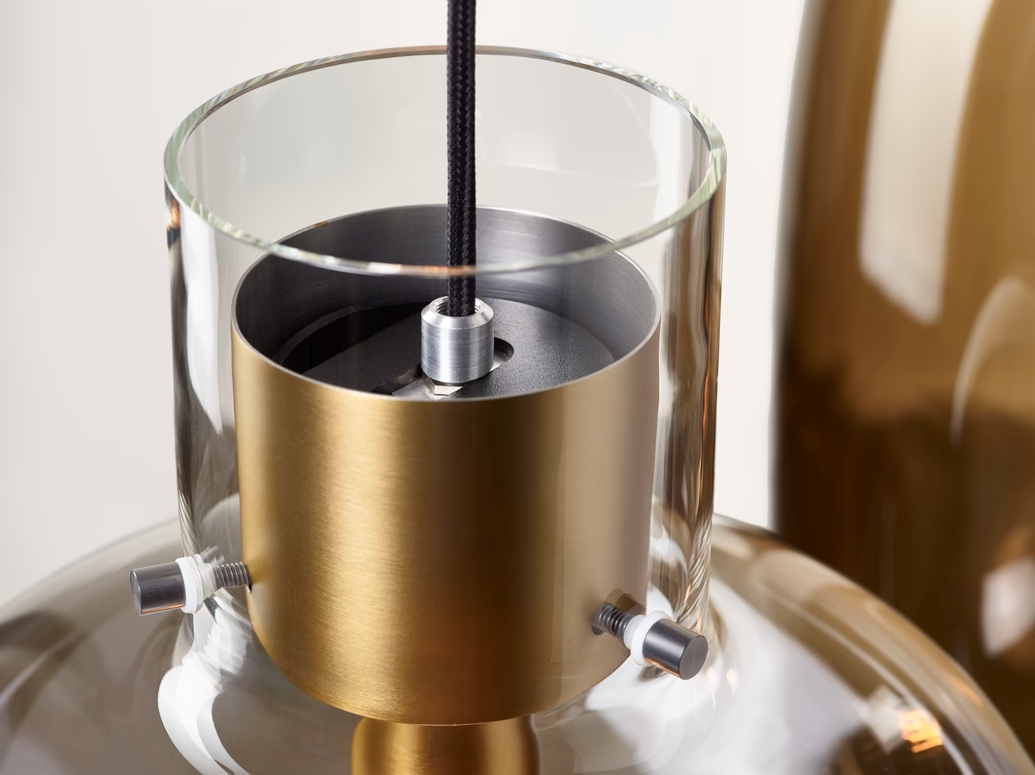 Close-up of Brokis Awa Pendant, handblown glass shade, brushed brass cylinder, luxe black flex; blurred twin behind.