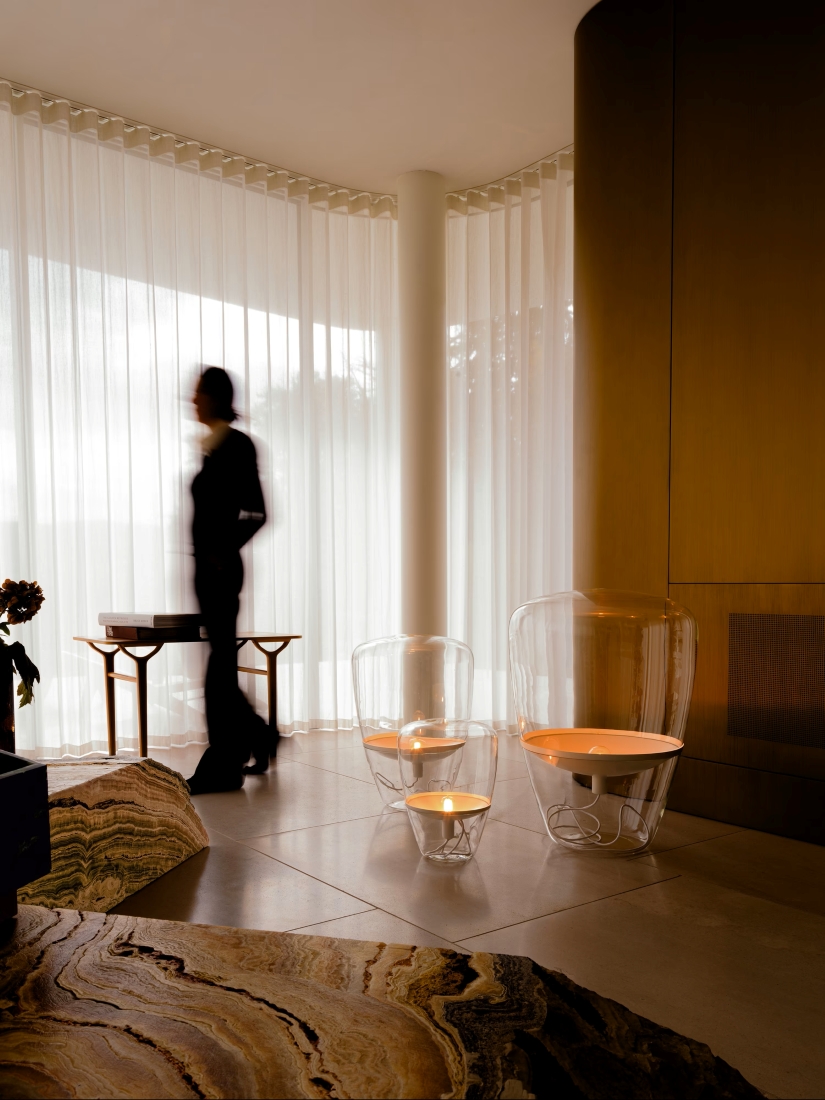 A blurred figure passes sheer curtains in a luminous modern room, with Brokis Balloons lamps near a golden-brown wall.