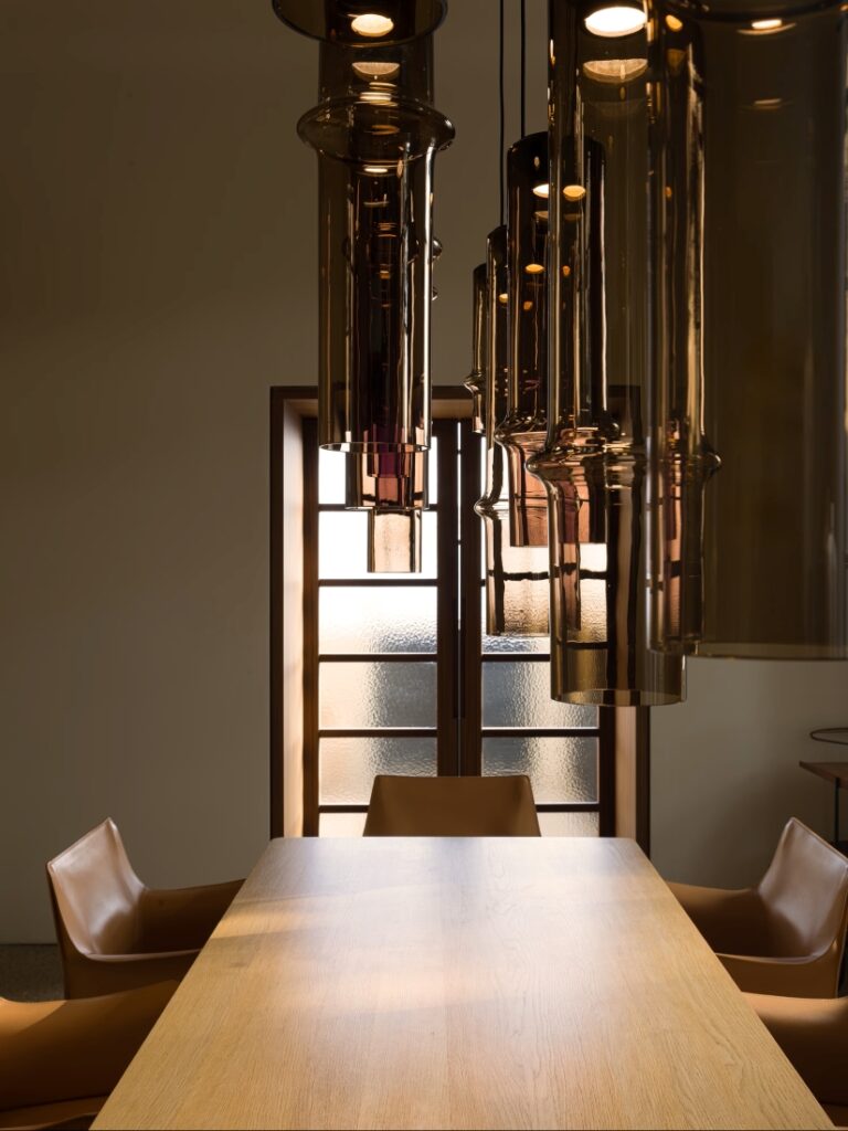 A modern dining room with a wooden table, tan leather chairs, and Brokis Bamboo Forest pendants overhead.