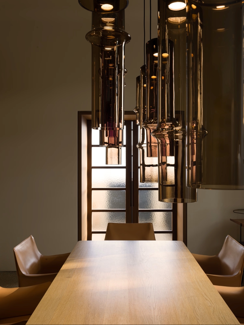 A modern dining room with a wooden table, tan leather chairs, and Brokis Bamboo Forest pendants overhead.