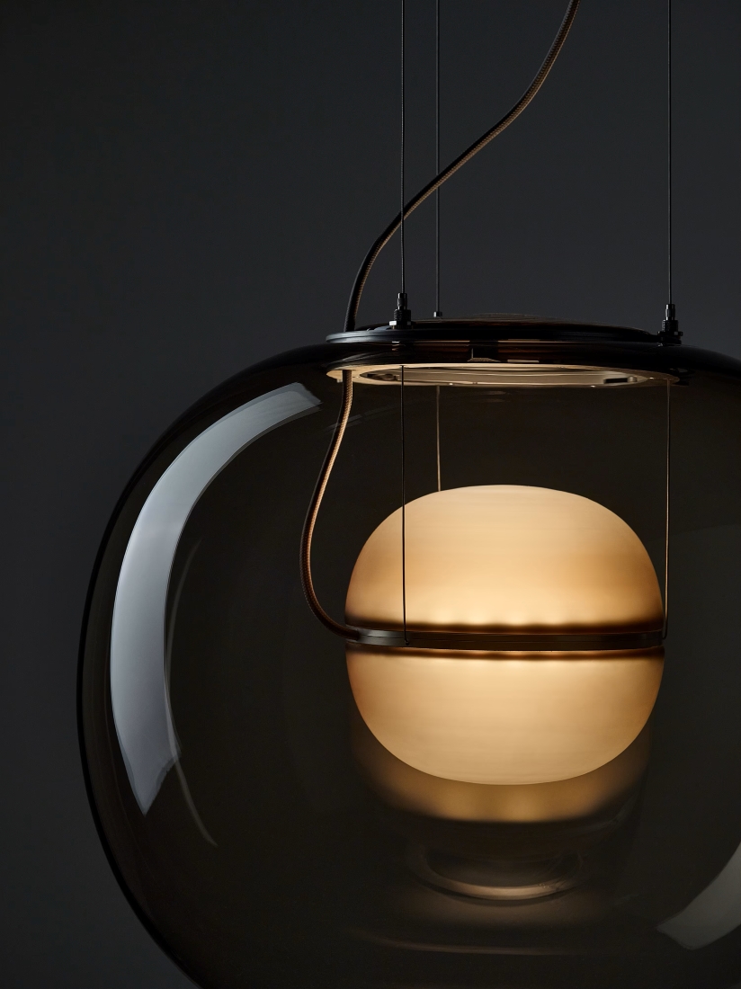 Brokis Big One Small Pendant with a glowing frosted orb, encased in clear glass, suspended by sleek rods against grey.