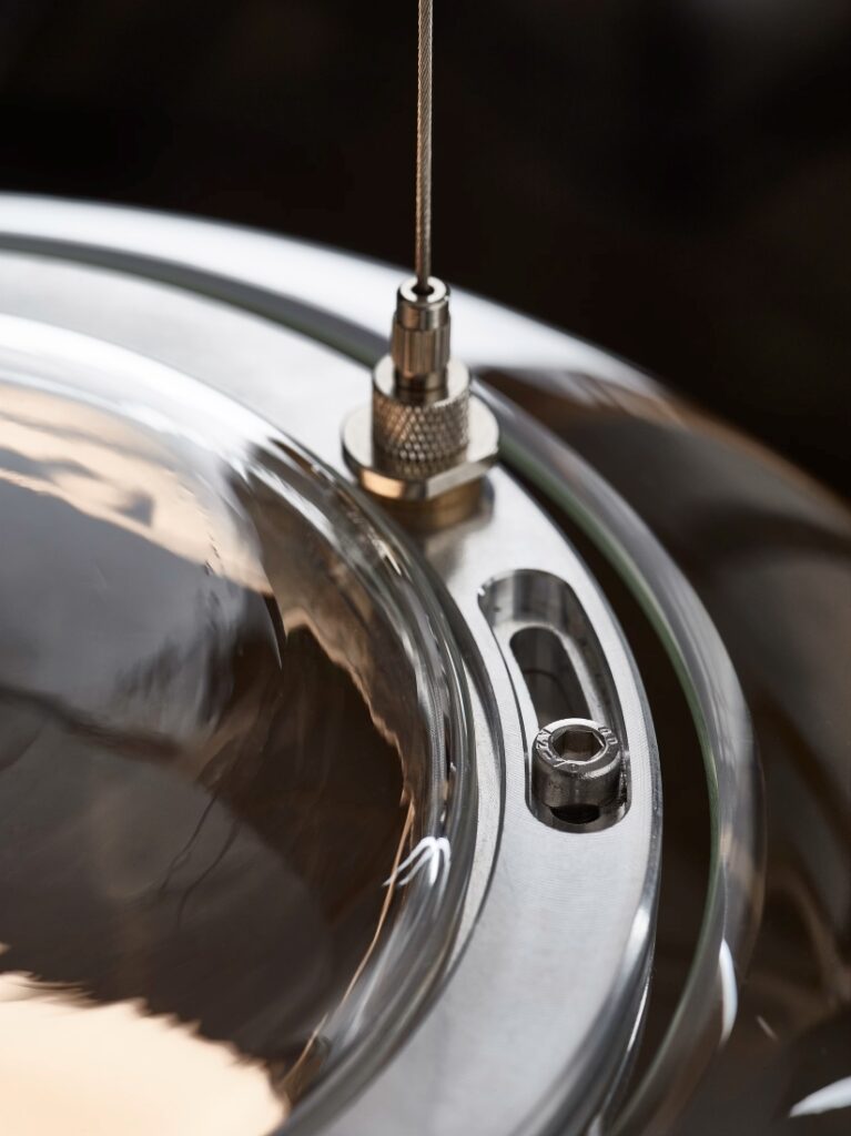 Close-up of a Brokis Big One metallic lid with round glass centre, cable fitting, and reflections on smooth metal.
