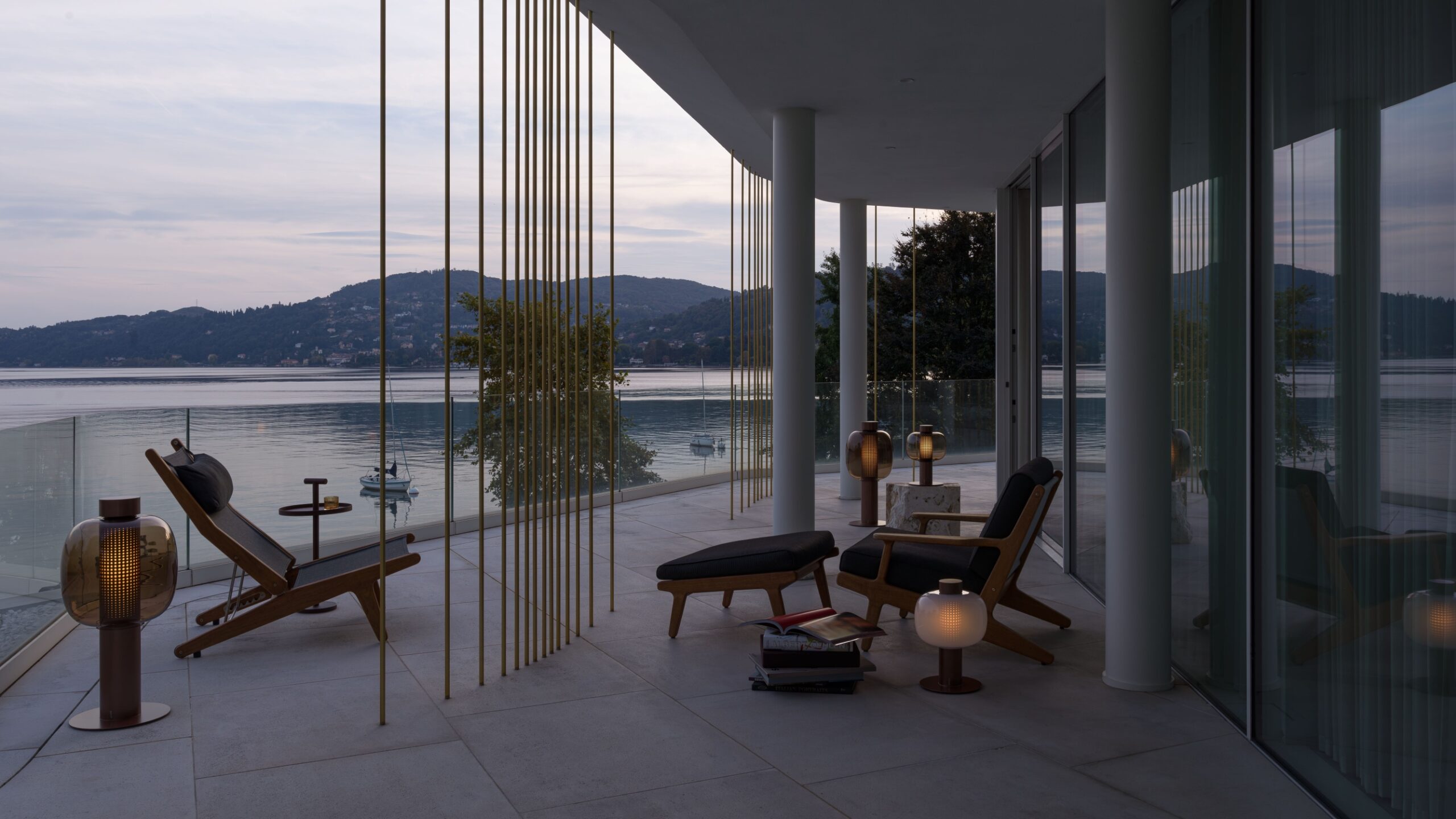 Brokis Bonbori outdoor lamps on a modern lakeside balcony at sunset, with wooden lounge chairs, by calm water.