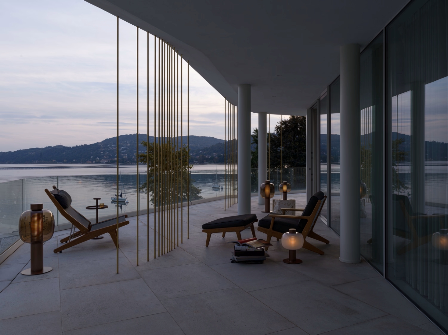 A modern lakeside terrace at dusk with Brokis Bonbori Outdoor lamps, gold railings, lounge chairs, and serene water views.