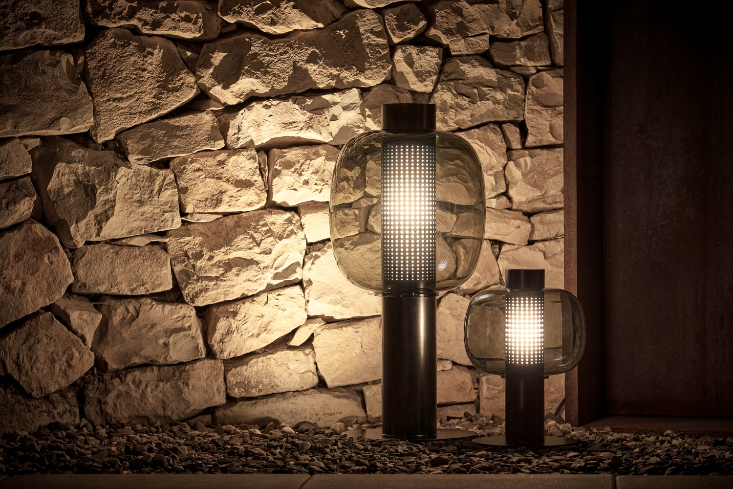 Sculptural Brokis Bonbori Outdoor lanterns with luminous metal bases cast a soft glow on a rustic stone wall at night.