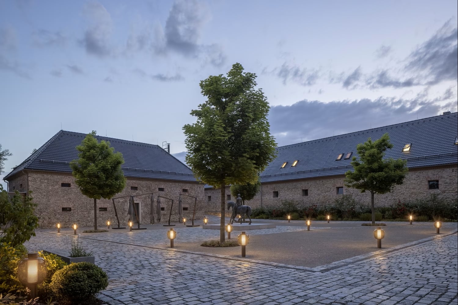 A cobblestone courtyard at dusk is lined with Brokis Bonbori Outdoor Large lanterns and manicured trees.