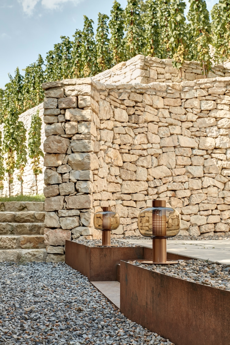 A modern garden with stacked stone walls, gravel paths, and Brokis Outdoor Small lanterns amidst lush green vines.