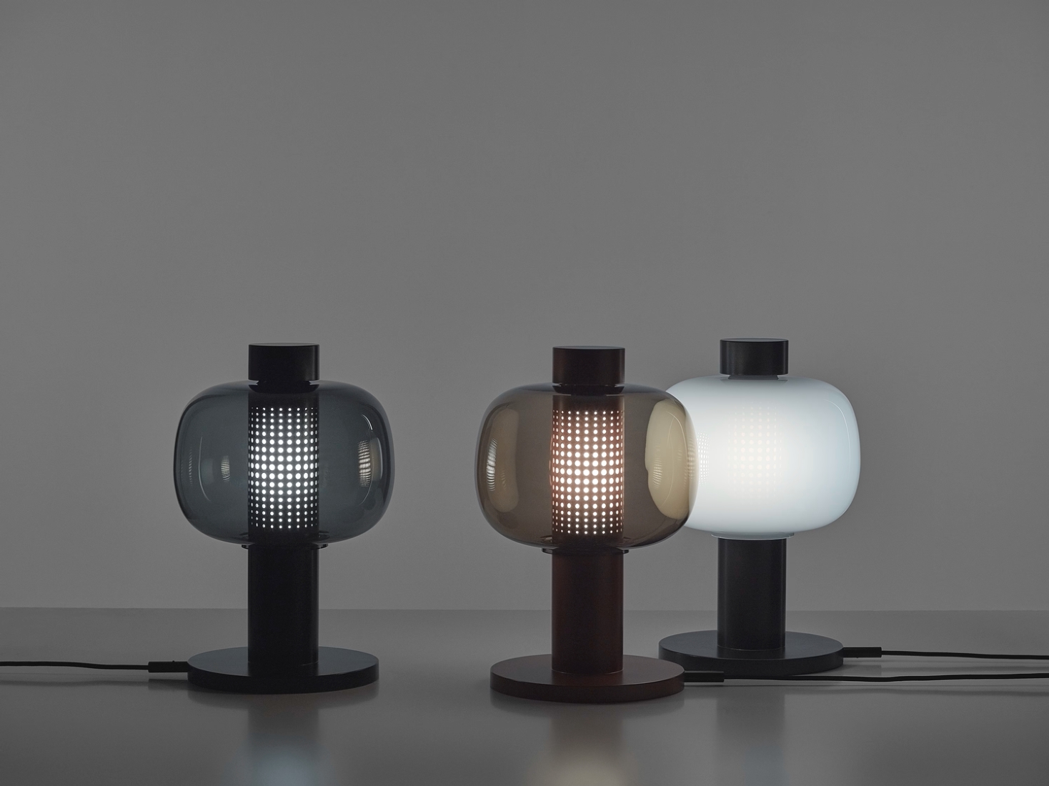 Three Brokis Bonbori Small table lamps with handblown glass shades emit a soft glow on a sleek grey surface.
