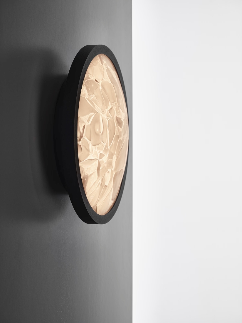 Brokis Convex M Applique wall light with handblown, BROKISGLASS emits a warm glow on a grey wall beside a white backdrop.