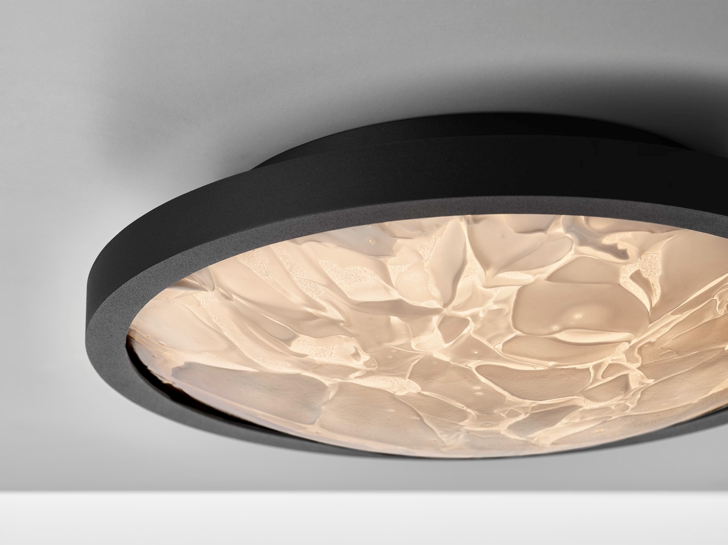 A close-up of a Brokis Convex M Applique ceiling light with a black frame and BROKISGLASS diffuser aglow on a white ceiling.