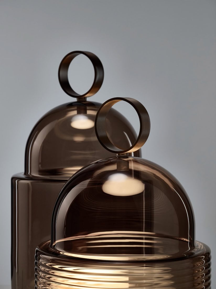 Two Brokis Dome Table lights with brown glass lids and bold handles, one ribbed, rest on an elegant light grey backdrop.