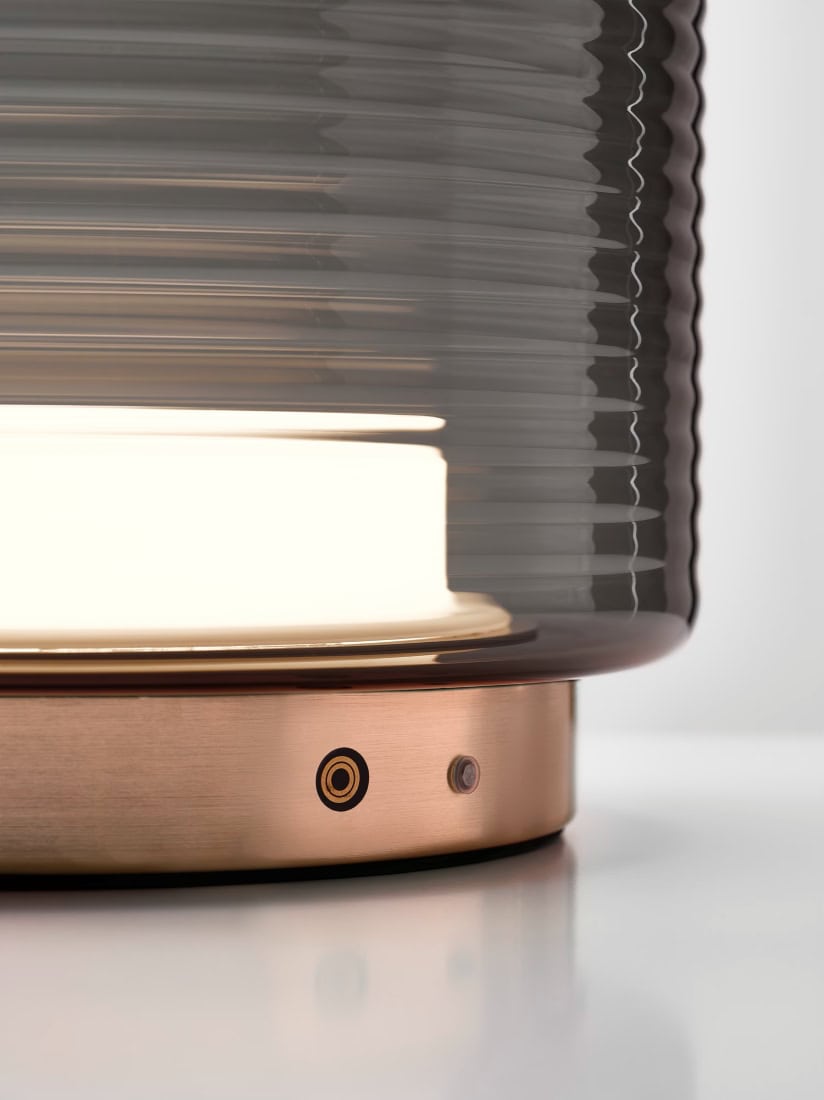 Close-up Brokis Dome Table lamp with ribbed glass shade, warm glow, and brushed copper base on glossy white surface.