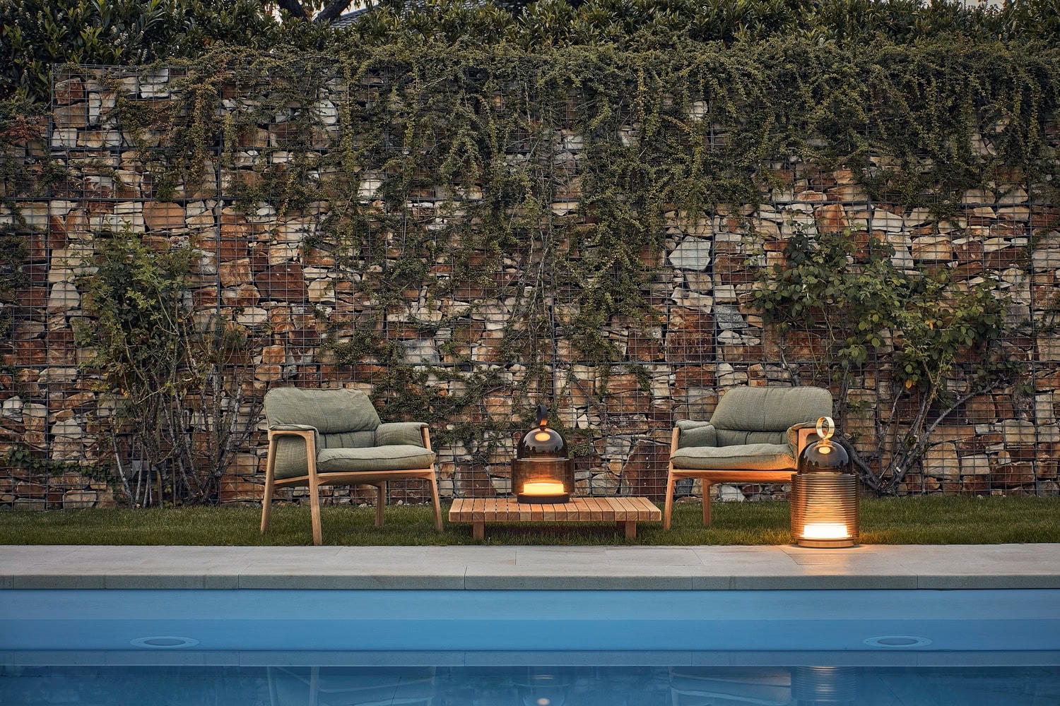 Elegant cushioned chairs and a low table sit poolside, lit by Brokis Dome Nomads; gabion wall greenery behind.