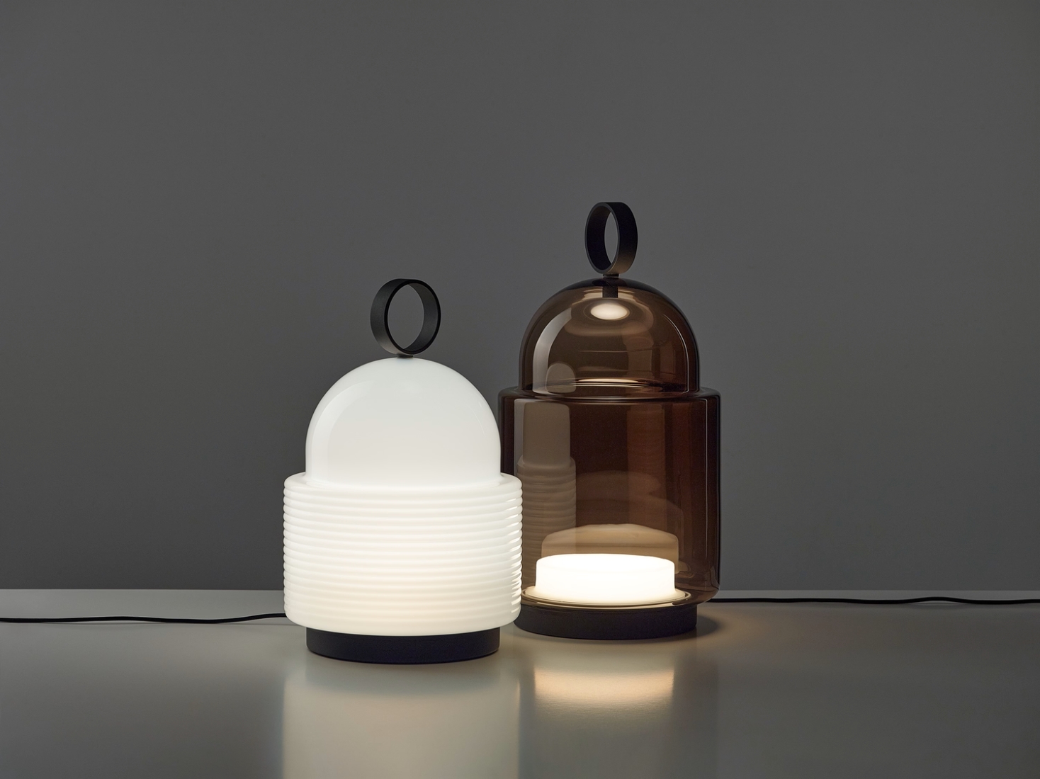 Two Brokis Dome table lamps with domed tops, handblown glass - one opaline ribbed, one smoke brown - on a grey surface.