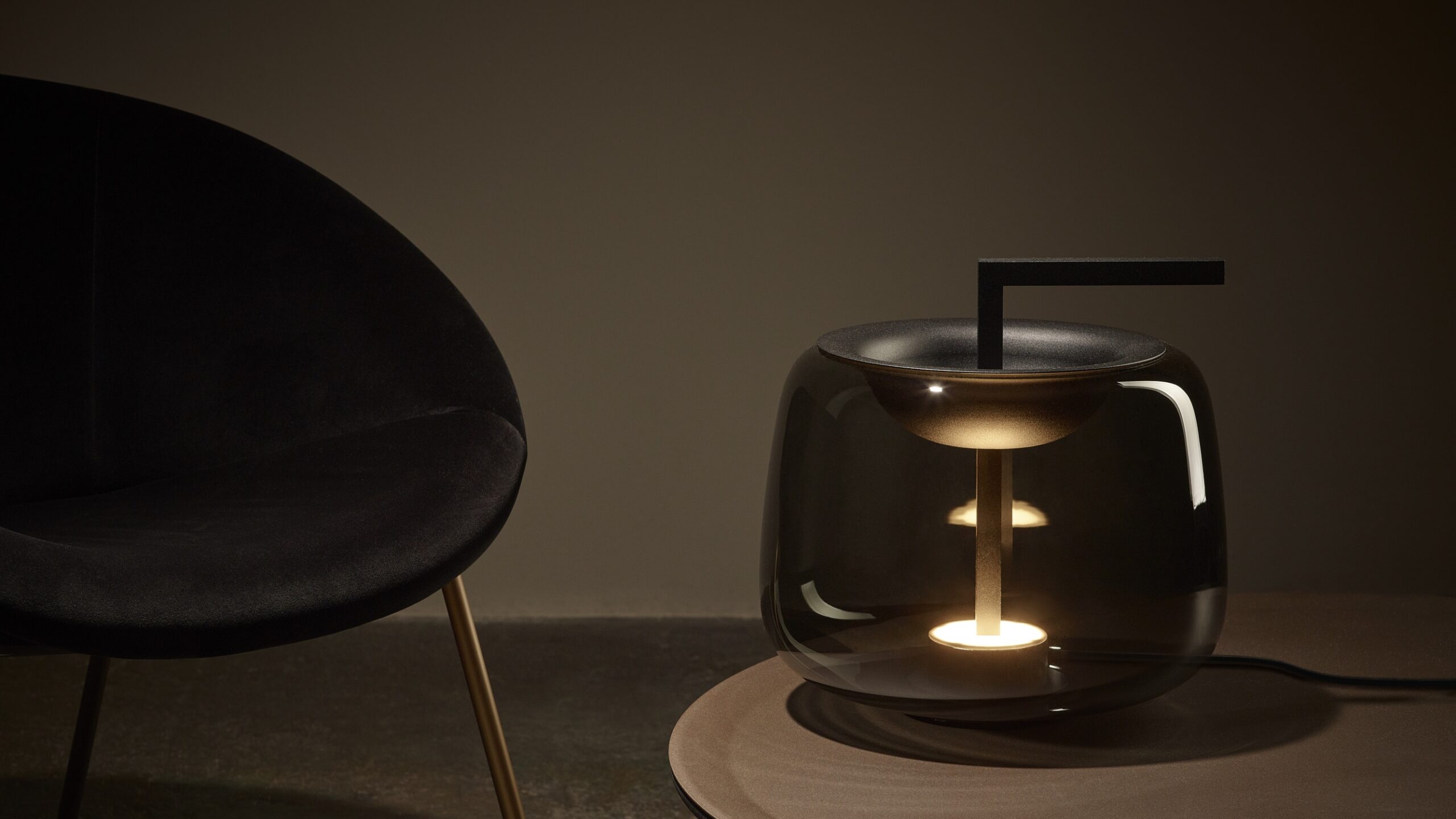 Brokis Double Table lamp with a grey glass base glows on a round table beside a luxe black chair with gold legs.