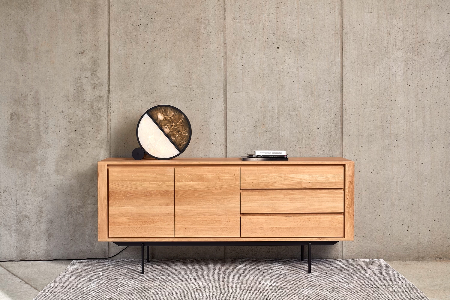 A light wood sideboard with black legs against concrete, topped with a Brokis Geometric Table lamp and curated books.