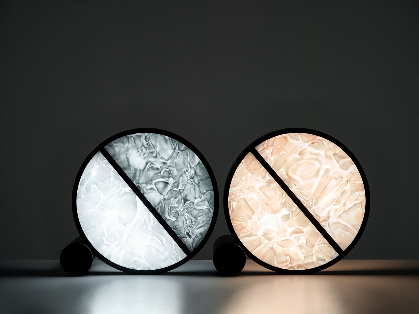 Two Brokis Geometric Table lamps—one cool white, one warm amber—shine on a smooth floor, framed by BROKISGLASS.