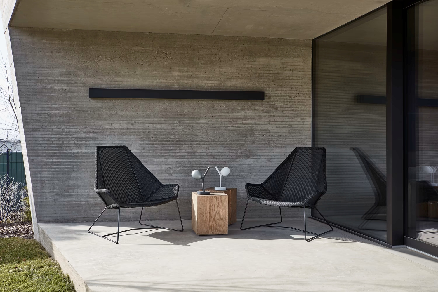 A chic patio set with two black woven chairs, a wooden cube table, and a Brokis Ivy Battery Table lamp exudes modern luxury.