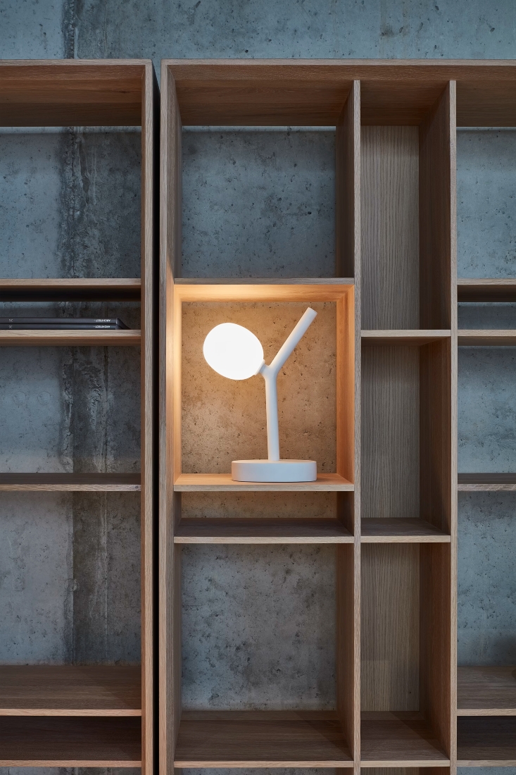 A white Brokis Ivy Battery Table lamp with a glowing orb accents a minimalist wooden shelf against a sleek concrete wall.