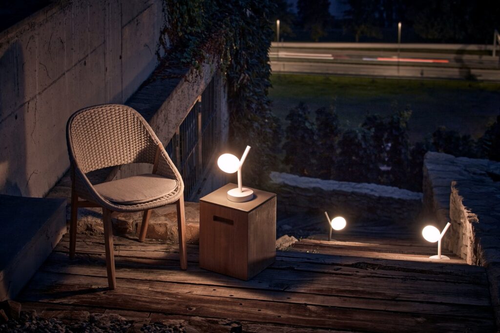 A luxe deck at dusk with a cushioned wicker chair, cube table, and  Brokis Ivy Battery Table lamps lining elegant steps.