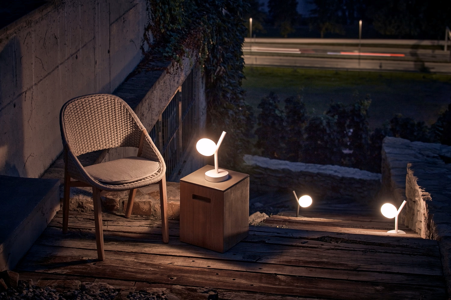 A luxe deck at dusk with a cushioned wicker chair, cube table, and  Brokis Ivy Battery Table lamps lining elegant steps.
