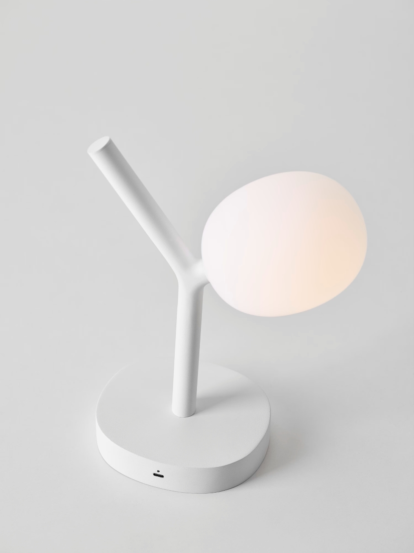A white Brokis Ivy Battery Table lamp with a frosted glass shade and USB port, glowing softly on a grey backdrop.